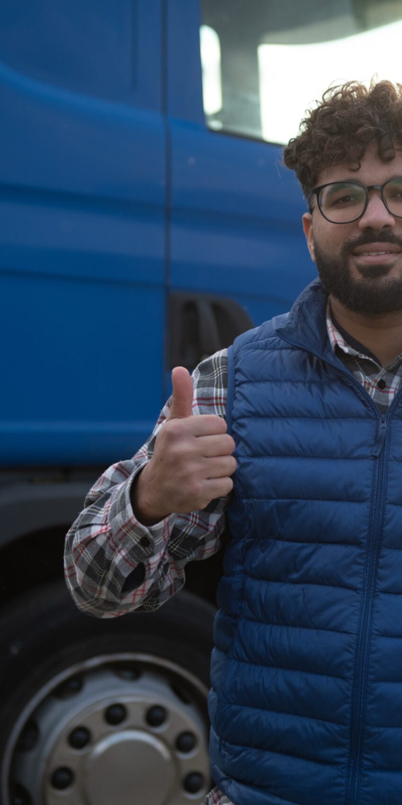 Happy man standing in front of a blue truck, smiling and showing a positive gesture about his job in logistics